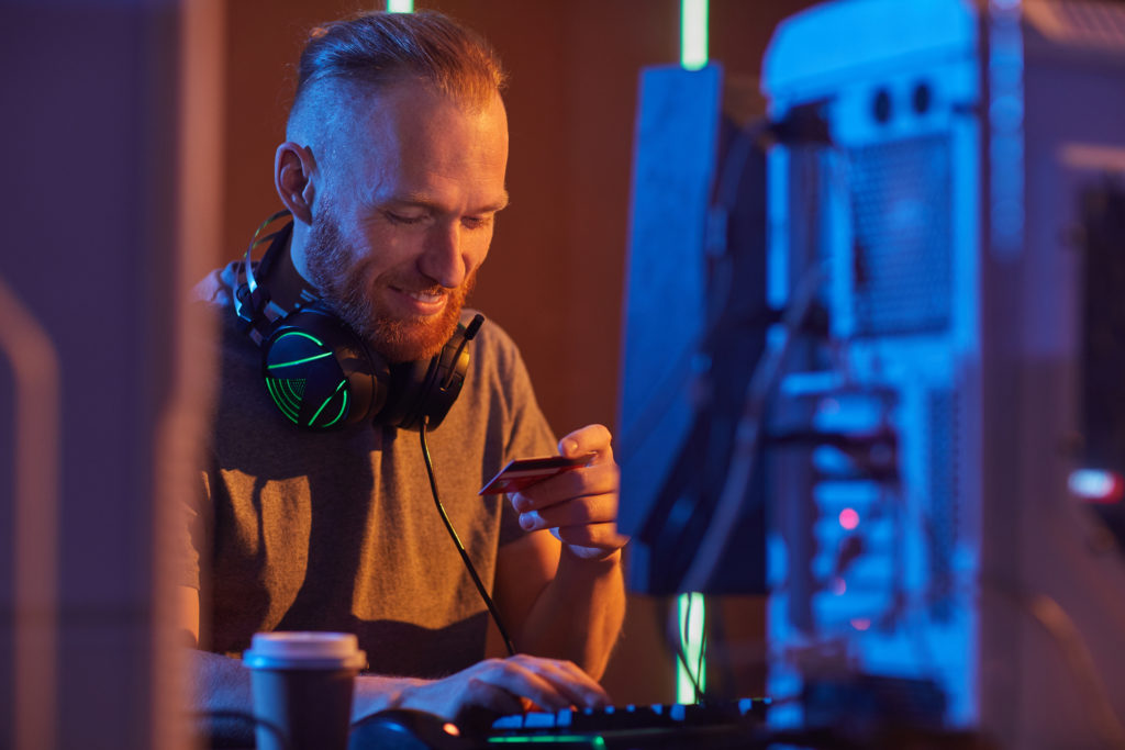 Young smiling man sitting at the table and paying online for computer game with credit card in dark room